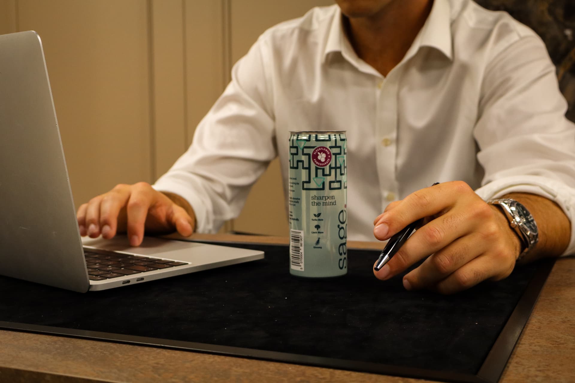 A can of sage on a desk beside a hand holding a pen and an open laptop